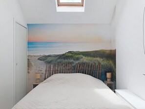 Une chambre avec une fresque murale représentant une plage, une lucarne, un lit et une table de chevet.