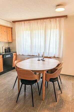 A dining area with a round table, wooden chairs, a kitchen with cabinets, and a window with sheer curtains. - Armeria by Interhome (Laax)