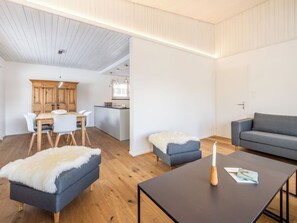 Ein modernes Wohnzimmer mit einer Couch, einem Hocker und einem Couchtisch, ein Essbereich mit Stühlen und Tisch sowie eine Küche mit Holzschränken und Pendelleuchten.
