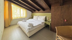 Ein Schlafzimmer mit Holzboden, einem Bett mit weißen Leinen, einem Nachttisch, einem Sessel und einem Fenster mit Vorhängen.