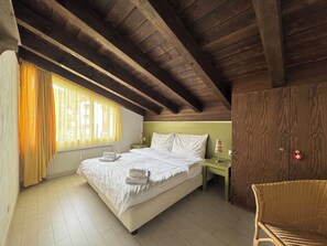 Ein Schlafzimmer mit Holzboden, einem Bett mit weißen Leinen, einem Nachttisch, einem Sessel und einem Fenster mit Vorhängen.