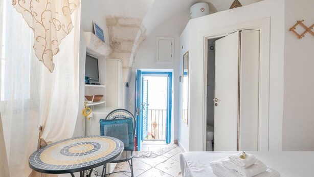 Ein schmales Zimmer mit einem Bett, einem Tisch und einem Stuhl. Eine Tür führt zu einem Balkon.