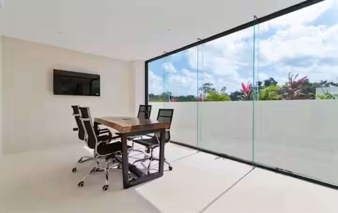 A modern room with a glass wall, a flat-screen TV, a wooden table, and office chairs.