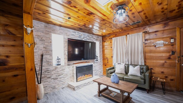 A cozy cabin interior with a stone fireplace, a leather sofa, a television mounted on the wall, and wooden paneling.