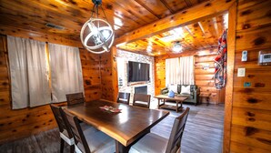 A wooden cabin interior with a dining table, chairs, a fireplace, and a television.
