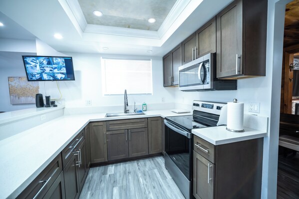 A modern kitchen with dark wooden cabinets, stainless steel appliances, a flat-screen TV mounted above the sink, and a window with blinds.