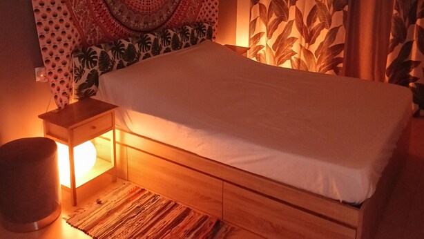 Ett sovrum med en säng, ett sängbord, en lampa, en väv och en gardin med ett lövmönster.