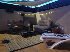 Un patio couvert avec une terrasse en bois, un banc rembourré, deux chaises, une petite table et des guirlandes lumineuses.