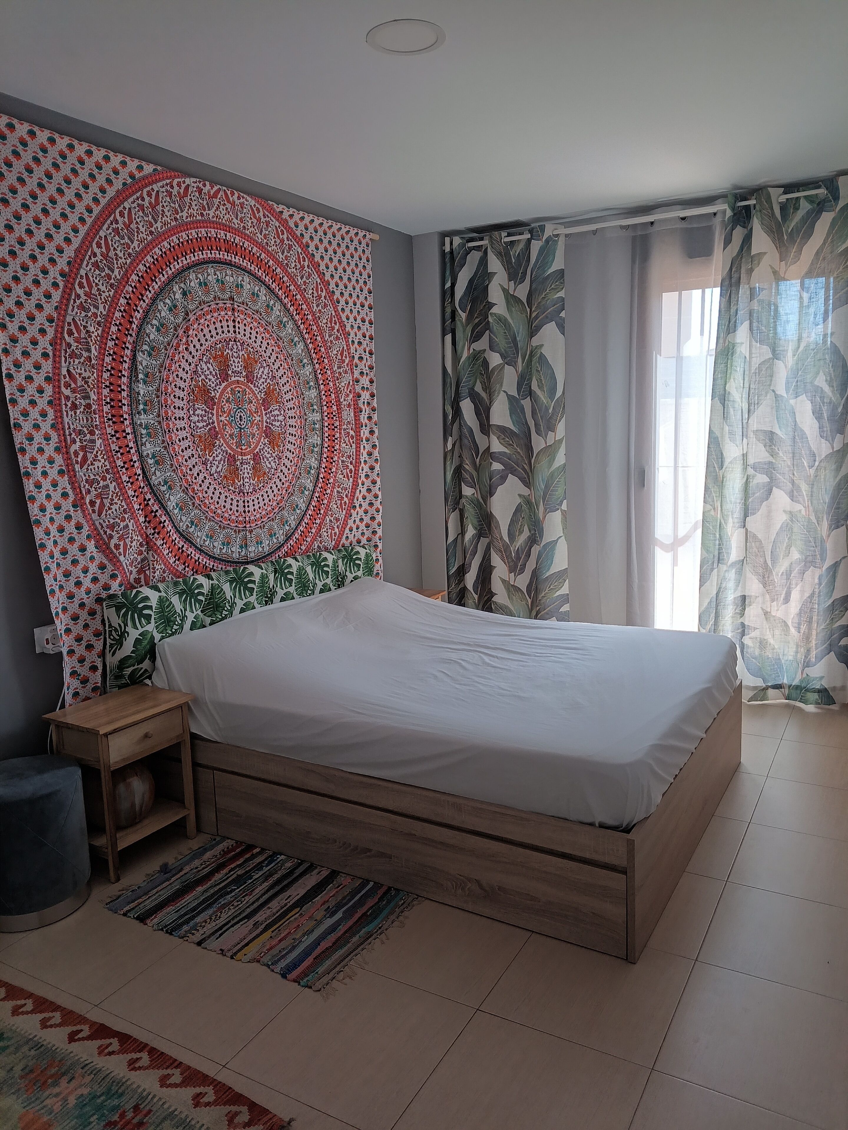 Une chambre à coucher avec un lit, une table de chevet, une tapisserie à motifs et une fenêtre avec des rideaux.