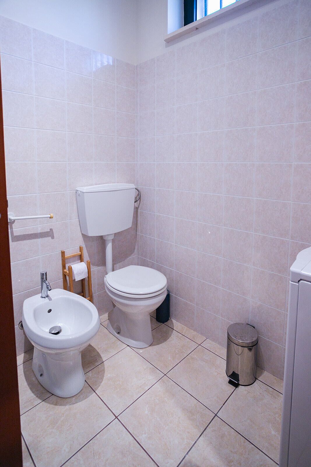 Une salle de bain comprenant des toilettes, un bidet et un lavabo, ainsi qu'un carrelage au sol et une petite poubelle.