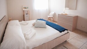 A neatly made bed with rolled towels, a bedside table with a lamp, a chair, and a dresser in a well-lit room. - Mediterranea - Seaside Sicily Home with Veranda (Marina di Modica (Modica))