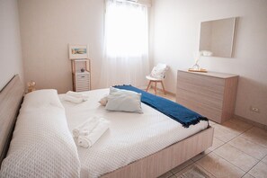 A neatly made bed with rolled towels, a bedside table with a lamp, a chair, and a dresser in a well-lit room. - Mediterranea - Seaside Sicily Home with Veranda (Marina di Modica (Modica))