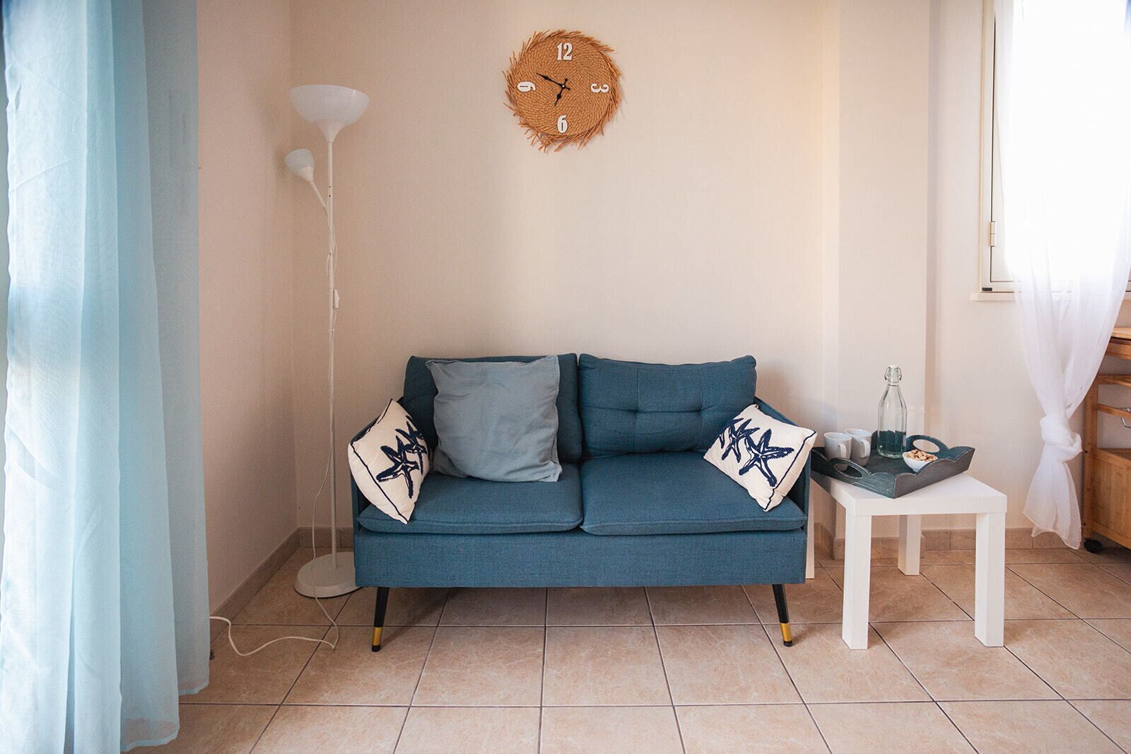 Una habitación con un sofá azul, dos almohadas, una mesita blanca con una botella y un cuenco, una lámpara de pie y un reloj de pared.
