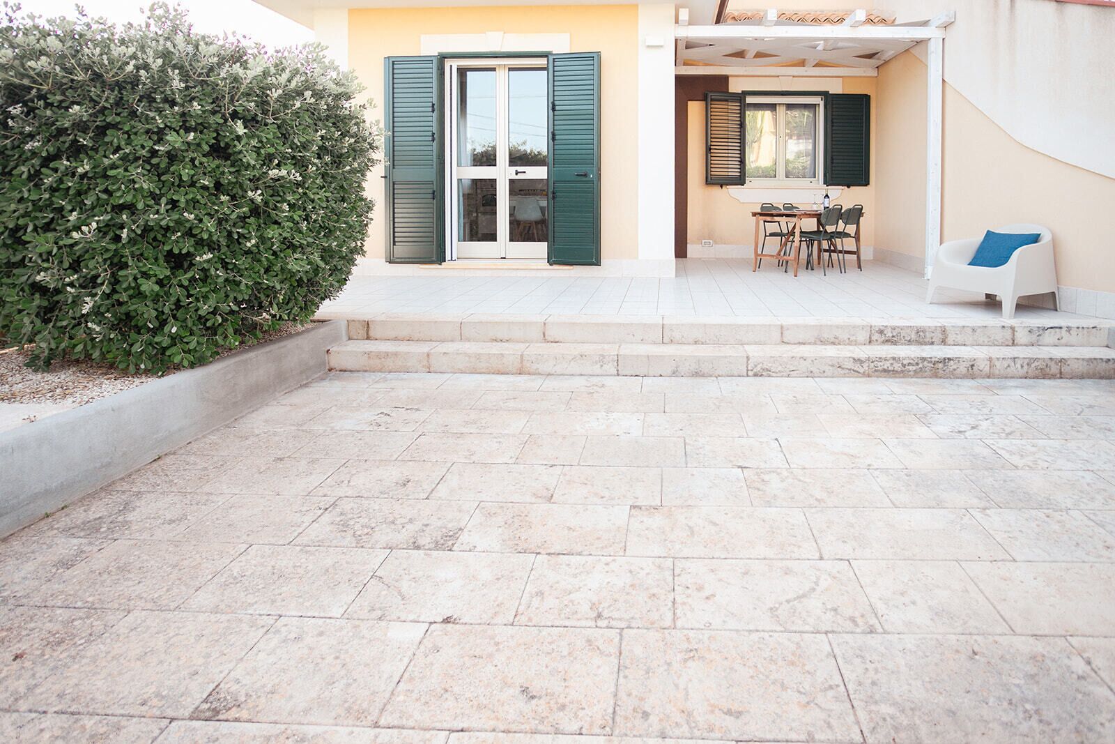 Un patio con suelo de baldosas, un seto verde, un cristal con contraventanas y una mesa con sillas.
