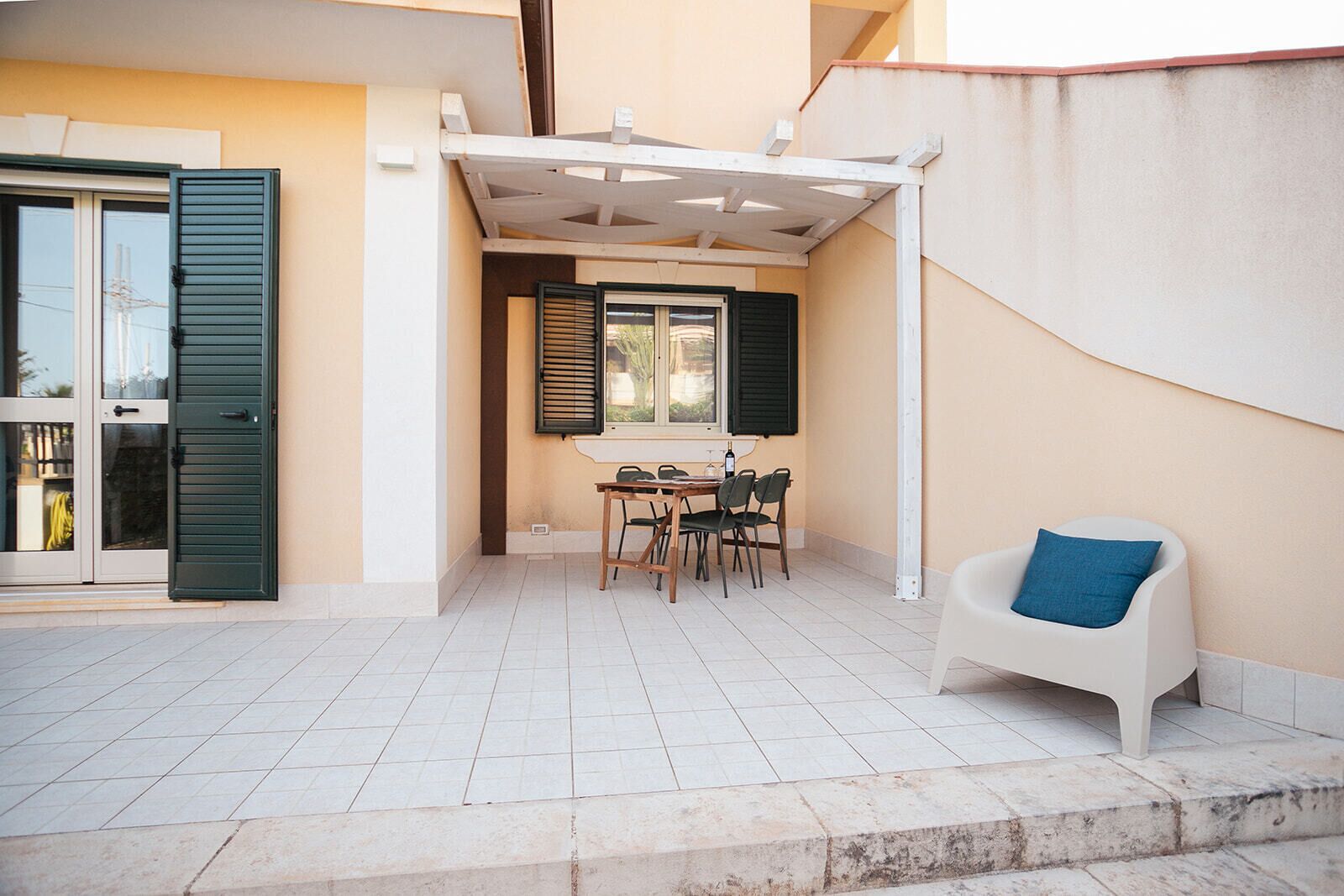 Un patio couvert avec une table et des chaises, une chaise blanche avec un coussin bleu et une fenêtre avec des volets.