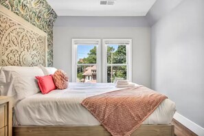 Une chambre à coucher avec un lit, deux fenêtres, une tête de lit à motifs et une vue sur l’extérieur.