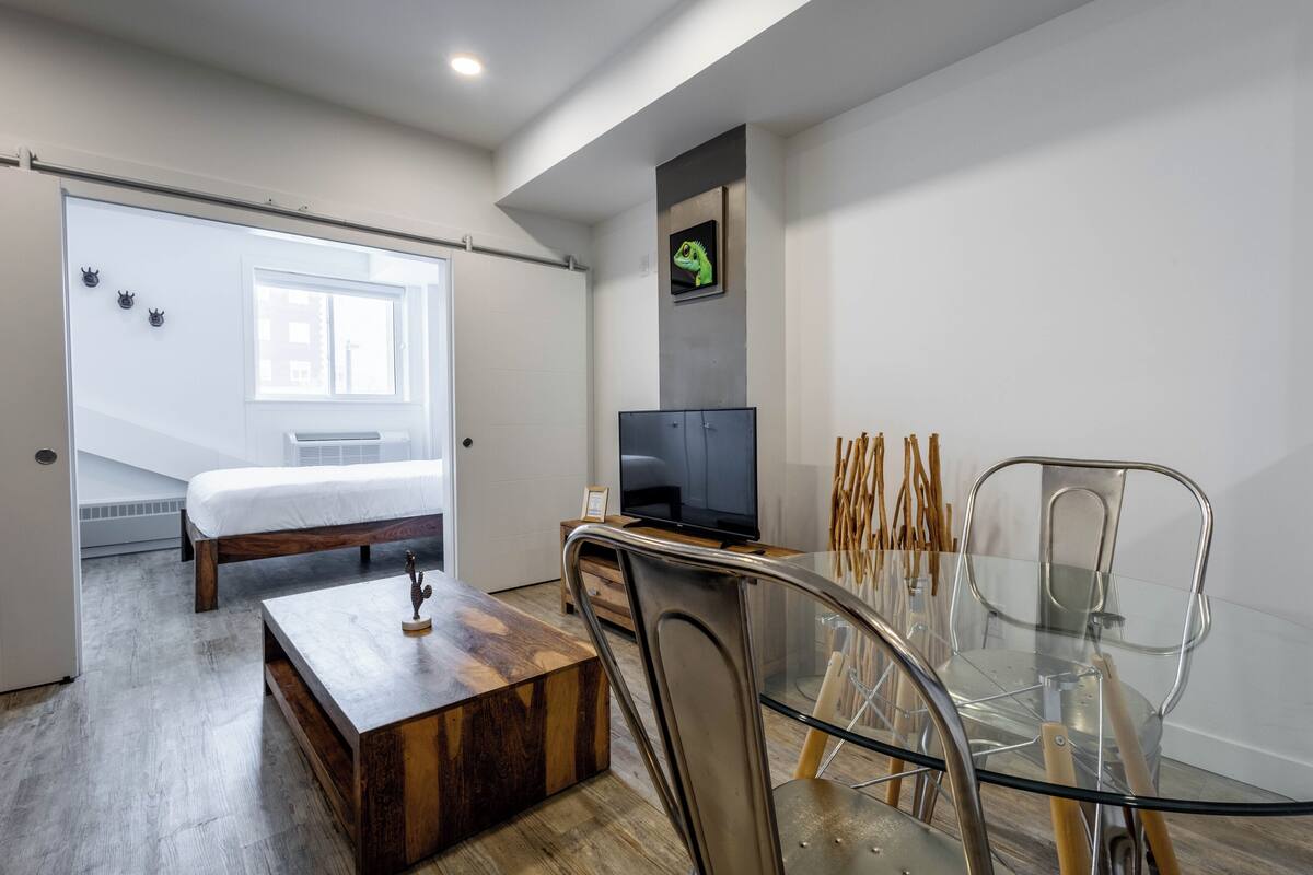 A modern bedroom with a bed, a TV, a glass dining table, and a chair.