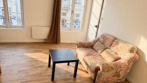 A room with a wooden floor, a floral sofa, a black table, and large windows with a view of buildings.