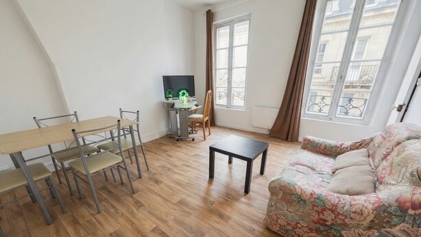A living room with a wooden floor, a sofa, a dining table with chairs, a TV, and a window with curtains.