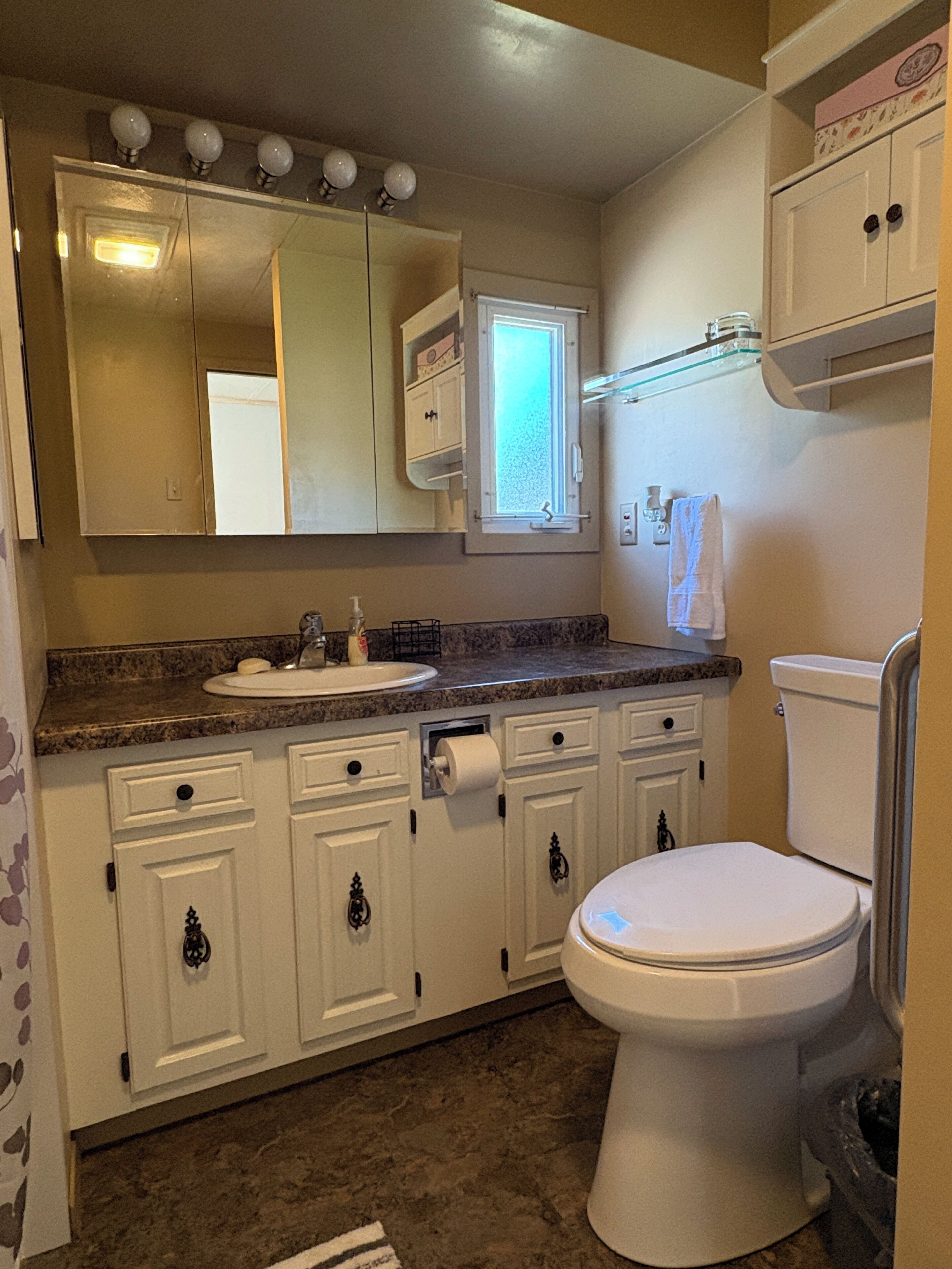 Une salle de bain avec des toilettes blanches, un lavabo avec un miroir au-dessus, et une fenêtre.