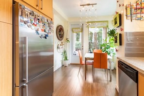 A modern kitchen with a stainless steel refrigerator, wooden cabinets, a dining table with orange chairs, and a view of the garden through the window.