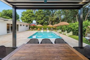 Ein überdachter Poolbereich im Freien mit einem Holztisch und -stühlen, umgeben von viel Grün und einem weißen Gebäude.