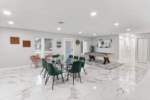 A spacious room with a glass-top dining table surrounded by chairs, a pool table, and a door leading to another room. - Villa Solstice | Heated Pool • Covered Terrace (Miami)