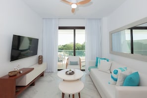 Ein modernes Wohnzimmer mit Flachbildfernseher, einer blauen Couch mit Kissen, einem runden Couchtisch und einem Balkon mit Aussicht.