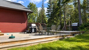 Een houten terras met tuinmeubilair, een barbecue en een rood gebouw in een bebost gebied.