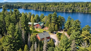 A house with a red exterior and a green roof, surrounded by dense forest and a lake.