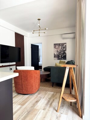 Uma sala de estar moderna com televisão de tela plana, uma poltrona redonda laranja, uma mesa de centro de madeira, um sofá verde e uma mesinha lateral de madeira.