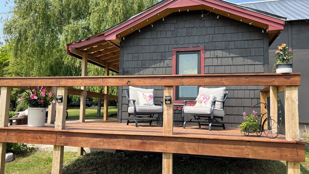 A wooden deck with two chairs and a table, a building with a dark exterior and a red roof, and a large tree in the background. - Perfect 1-bedroom  off barn with AC in charming Petoskey (Petoskey)