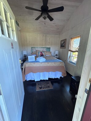 Un dormitorio con cama, ventilador de techo, ventana, puerta y felpudo.