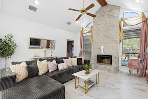 A modern living room with a sectional sofa, a fireplace, a ceiling fan, and a large window with curtains. - Velvet Paradise-6 bdrm estate with pool, hot tub, tiki bar and firepit (Mesquite)