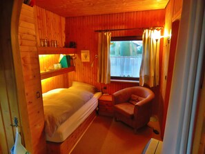 A cabin room with a bunk bed, a chair, a window with curtains, and a shelf with items. - The perfect place to do nothing | A wooden house nestled between the forest and meadows (Geestland)