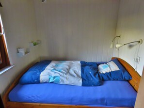 Une petite chambre avec un lit simple, une tête de lit en bois, une couvre-lit bleue, un mur blanc et une lampe de chevet.