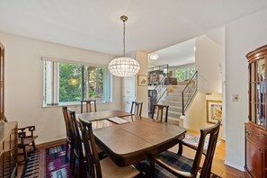 A dining area with a table and chairs, a chandelier, a staircase, and a view of greenery outside.