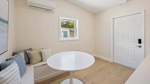 Una habitación con un equipo de aire acondicionado blanco, una ventana pequeña con vistas a vegetación, una puerta blanca y una mesa redonda blanca.