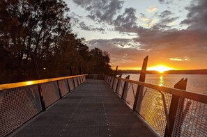 A pedestrian bridge with a metal railing, leading to a body of water during sunset. - Private Lower‑Level 2‑Bedroom Unit – Walk to Lake Macquarie (Eleebana)