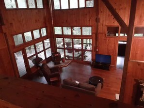 A wooden cabin interior with large windows, a fireplace, a sofa, a television, and a dining area.