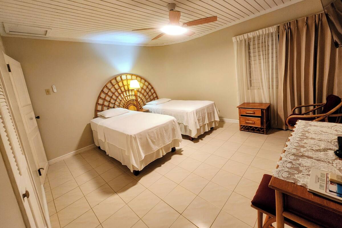 Une chambre d’hôtel avec deux lits, une tête de lit en bois, un bureau avec une chaise, une fenêtre avec des rideaux et un ventilateur de plafond.