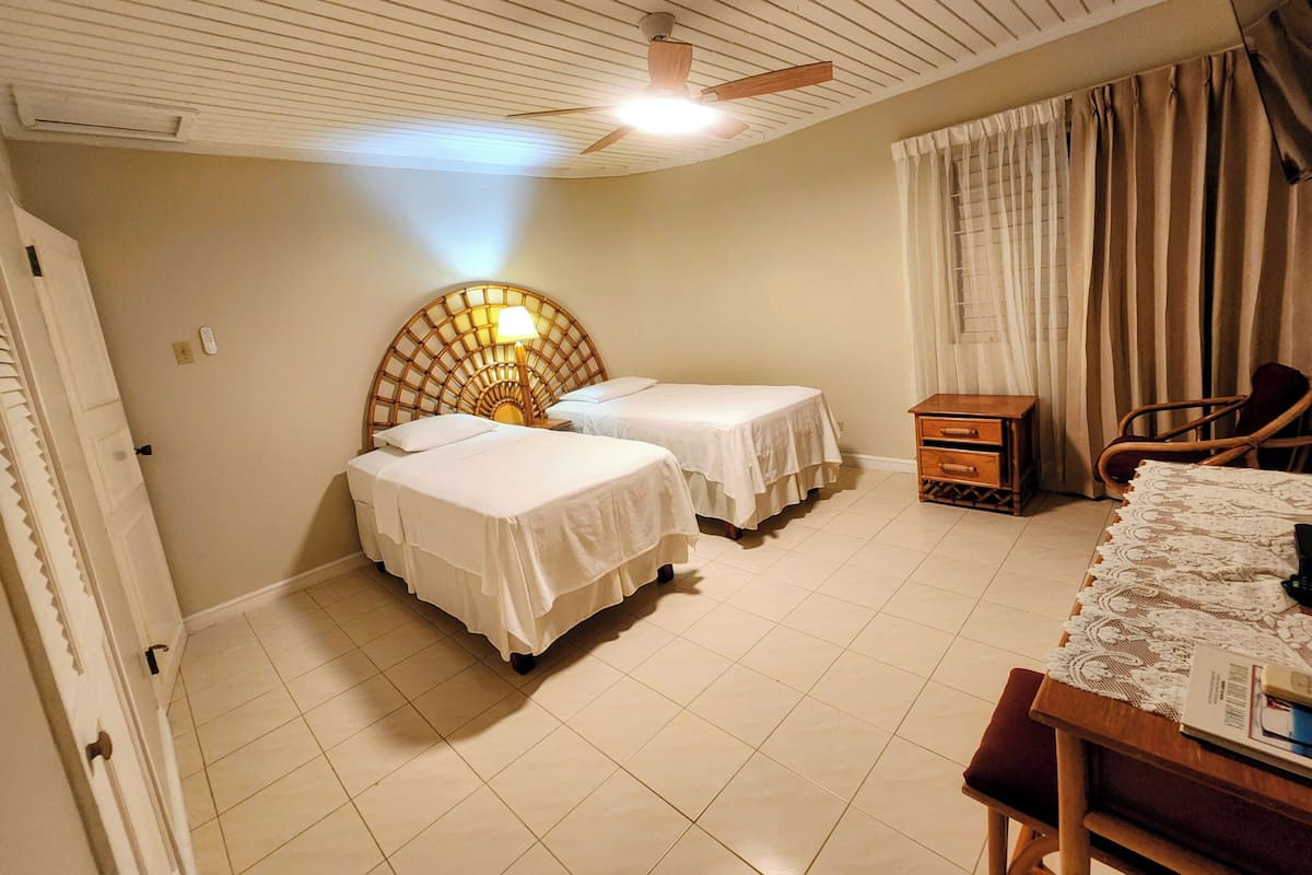 A hotel room with two beds, a wooden headboard, a desk with a chair, a window with curtains, and a ceiling fan.