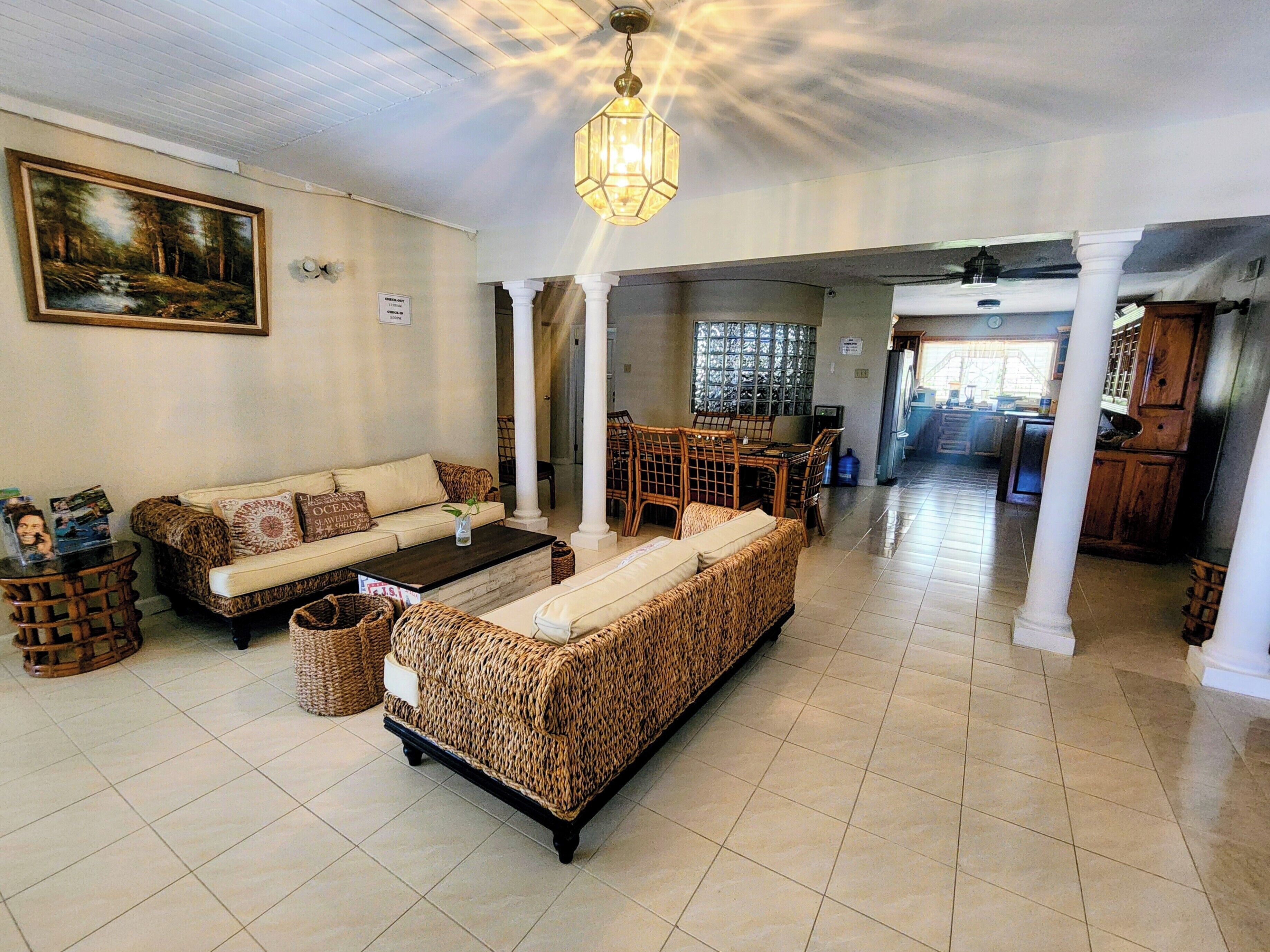 A spacious living area with a sofa, coffee table, and wicker furniture. There is a dining area with a table and chairs, and a kitchen in the background.