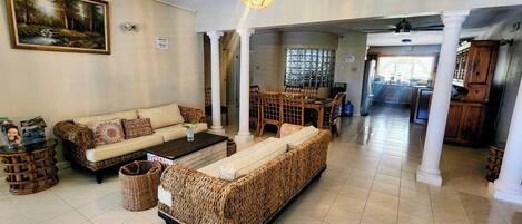 A spacious living area with a sofa, coffee table, and wicker furniture. There is a dining area with a table and chairs, and a kitchen in the background.