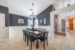 Een eetruimte met een donkere wand, witte lambrisering, een kroonluchter en een houten eettafel met stoelen.