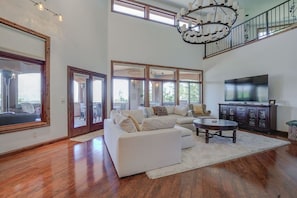 A spacious living room with a large white sectional sofa, a coffee table, and a TV on a dark wooden cabinet. There are large windows providing natural light and a view of the outdoors.