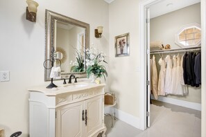 Un baño con un espejo grande, tocador y un vestidor con ropa colgada.
