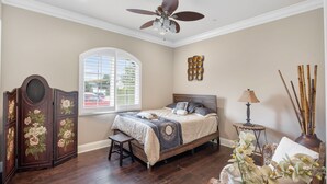 Un dormitorio con cama, ventilador de techo, ventana con persianas, un mueble decorativo y una mesita de noche con lámpara.