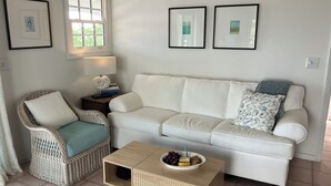 Een woonkamer met een witte bank, een rieten stoel en een kleine houten tafel met een schaal fruit.