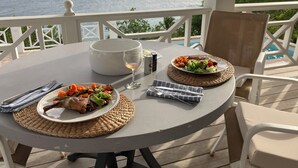 Een ronde tafel met borden eten, een glas wijn en een witte schaal op een balkon met uitzicht op zee.